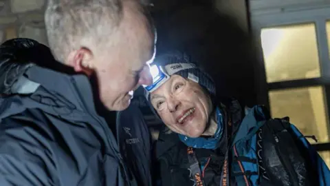 The Montane Spine Race Runner Anna Troup and her husband at the finish line of the Spine Race. Anna is wearing navy-coloured fell running gear, a head torch and a woolly hat. Her husband is also wearing a navy waterproof jacket.