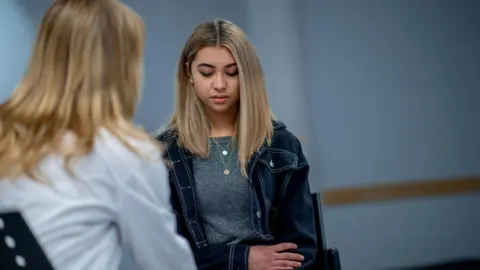 A teenage girl with long blonde hair and wearing a denim jacket and grey T-shirt has her head slightly bowed as she speaks to a woman with long blonde hair and a white shirt who is facing away from the camera.