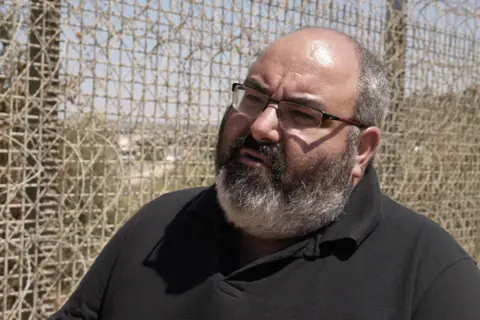 Oren Rosenfeld/BBC Yehuda Shaul wearing a black Polo shirt, bearded and with glasses in front of a tall fence