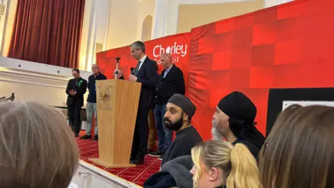 A man in a suit stands speaking at a lecturn on a stage. It has a sign on the front of it which reads 'Chorley Council'. Other men stand on the stage and others mill around below it. 