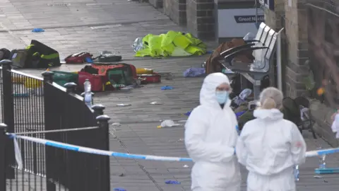 PA Media Two emergency service workers in forensic suits stand in the right hand corner of the image. Police cordon has segregated an area where paramedic equipment, clothing and other belongings lie on the floor