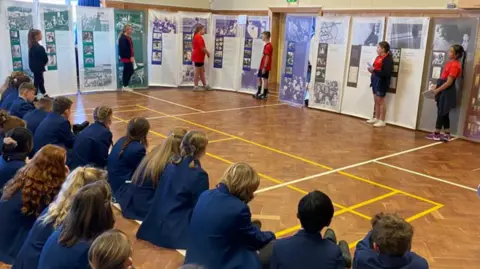 Simone Beach Children in blue blazers sitting on a brown wooden floor with their backs to the camera watching pupils share artwork and photographs from a history lesson.