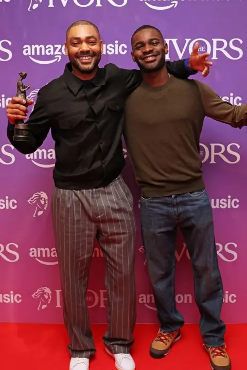 Getty Images Kano and Dave smile and stand on the Ivors red carpet. Kano holds his Ivor Novello Award.