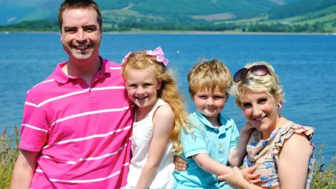 Susan, Lily, Hamish and Susan pictured on holiday. A large area of water behind them is blue and there are green, tree-covered hills on the opposite shore.