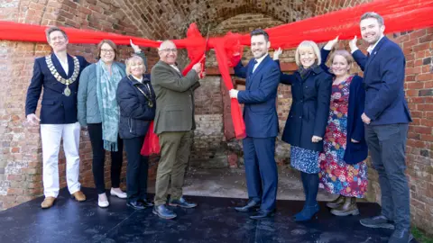 NWLDC Dignitaries using a giant pair of scissors to cut a big red ribbon tied around the furnace