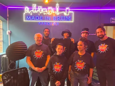 Students stand under neon sign of skyline outline and the phrase ‘Made in Brum Studios’ in a recording studio with their teachers and two rappers