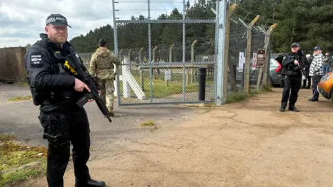 Matt Precey/BBC Two police officers with guns standing guard by a gate which is being opened by a US airman. They are dressed in black. The officer in the foreground has a beard.