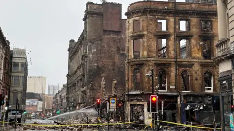 The fire-ruined blackened remains of a sandstone building on the corner of a junction in Glasgow city centre. The scene is cordoned off with yellow tape and the building's rubble surrounds the building perimeter.