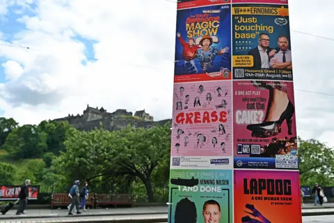 Ken Jack/Getty Images Brightly coloured posters advertising shows are pasted on a board with Edinburgh Castle in the background and the green trees of Princes Street Gardens below.