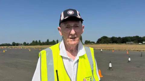 BBC/Emily Johnson A man in a hi-vis vest stands on tarmac at an airfield. The tarmac is lined with orange cones.