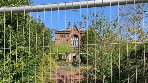 An old sandstone building viewed through fencing with police tape in front of it and some overgrown bushes
