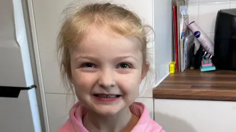 Maisie, five, smiles at the camera. She has blonde hair up in a ponytail. She has brown eyes and is wearing a pink hooded top.