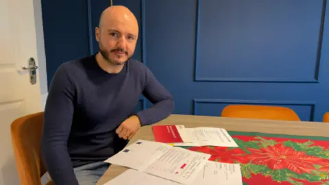 BBC Sevak Maljian sat at a table with various papers in front of him. He is bald with a dark beard, wearing a navy jumper.