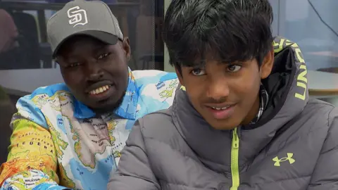 BBC A man and a boy sat smiling, one in a grey cap and a shirt with a world-map design on it, and the other in a grey and green coat.