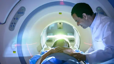 Image of a woman in blue hospital gown at the entrance to a white MRI/CT scanner lying face up at a male member of medical staff, dressed in a white coat.