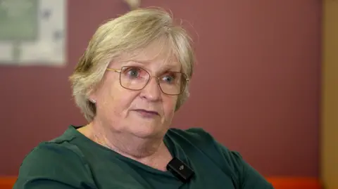An older woman with short blonde hair is seated indoors, wearing a green top with a small microphone clipped to the collar. She is wearing glasses. The background is softly blurred, featuring a purple wall and indistinct objects.