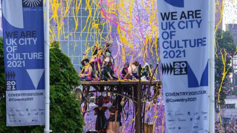 PA Media UK City of Culture 2021 banners fly in the foreground of an image of acrobats surrounded by colourful paper streamers which fill the sky.