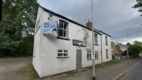 The building appears to be vacant and somewhat neglected, with several signs of disrepair visible on the exterior. A large "FOR SALE" sign is attached to the upper corner of the building. Above the former entrance is a faded hanging sign. Another small hanging sign is mounted higher up on the façade. The central doorway at the front is boarded up with plywood, and some of the signage appears weathered and damaged. The building has white-rendered walls with black trim around the windows and roofline. Several windows are visible on both floors. Some windows appear intact, while others look aged. A chimney rises from the roof near the centre of the structure.