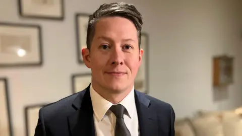 A man in a black suit jacket, black tie and white shirt. He has dark hair, combed to the side. He is smiling at the camera, standing in a room with cream sofas and black framed prints on grey walls.