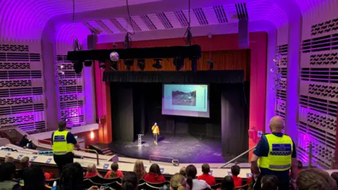 West Northamptonshire Council Interior of a theatre, showing a person in a yellow shirt and dark trousers walking on stage near a small lectern, and an image projected onto the back of the stage. There are red curtains and boxes on both sides with a wavy line pattern on the walls. Children sit in the theater seats, with a wooden plank in front of them. At the front of the seating area are two police officers in yellow hi-viz.