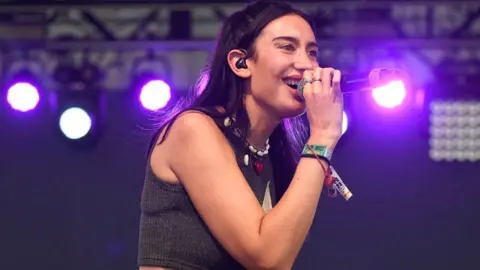 Julia Wolf sings on stage in a grey tank top and beaded necklace in Austin, Texas. She has dark hair.