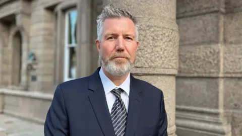 Adrian Moss is pictured in Jersey's Royal Square. He has a navy blazer on and a stripey tie. His hair is short, grey and spiky. He has a medium length beard. 