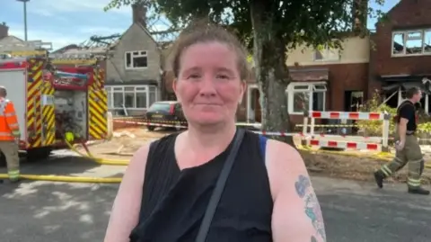 A woman with brown hair tied back is wearing a black vest and has tattoos on her left arm. She is standing in front of a house that has no roof
