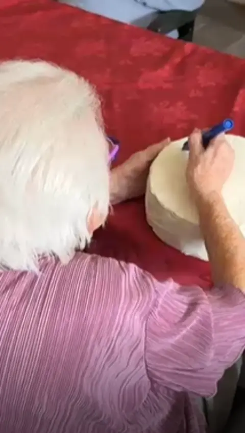 An elderly woman wearing a pink top is using a marker to write on a large wheel of cheese.