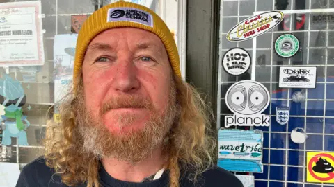 BBC/Richard Edwards A close-up portrait of Crawford standing outdoors in front of a storefront window. He is positioned centrally in the frame and is looking directly toward the camera. He has long, curly hair, a full beard, and several long hair strands hanging down in front of the shoulders. A mustard-yellow knitted beanie is being worn, featuring a rectangular patch with the text "SURFERS AGAINST SEWAGE" on the front.