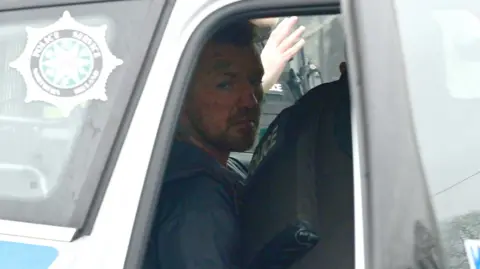 Pacemaker Man sitting in the back of a police car with an officer leaning over him