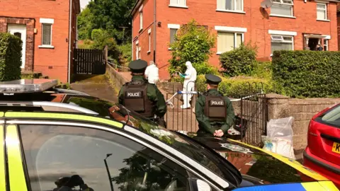 A photo taken from the opposite side of a road showing a parked police car outside a row of semi-detached, red brick houses in north Belfast. Three uniformed officers are standing with their backs to the camera. A red car is also parked blocking the driveway of the house. A cordon is placed along the bottom and mid-way up the driveway of the house.