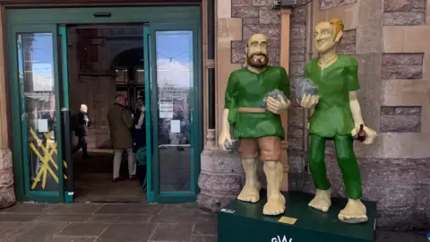Two giants wearing green outfits are appearing as a sculpture, standing next to the entrance of a train station's sliding doors.