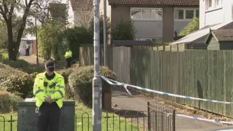 A footpath by some modern houses. There are trees and a grassy area on the left. Police tape has sealed off the path and three officers in hi-vis yellow vests can be seen at stages into the distance. It is sunny.