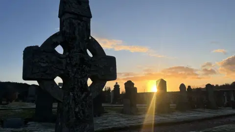 A photo of the cemetery at sunset. The sun is low and shines bright orange on the dark stone graves. These are the old graves not the new ones.