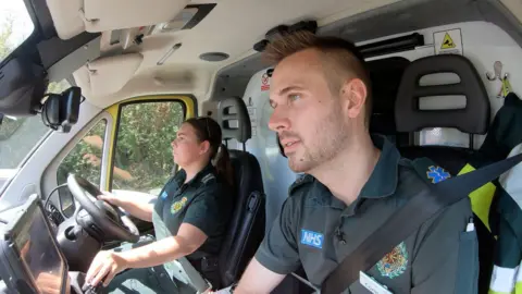 Two paramedics in their ambulance, dressed in dark green polo shirts with the NHS and ambulance service logos