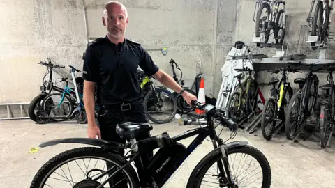 Un bărbat chel privește direct în cameră. Poartă un tricou negru și pantaloni scurți și ține în mână o bicicletă mare și neagră. În spatele lui sunt alte biciclete rezemate de perete și aliniate pe suporturi.