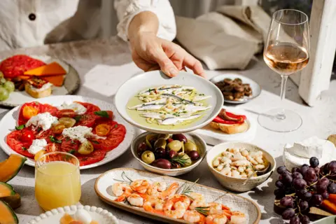 Getty Images A hand serving a plate of marinated anchovies over a table filled with Mediterranean mezze including shrimp, olives, tomatoes, cheese, beans, grapes, melon, and a glass of white wine. 