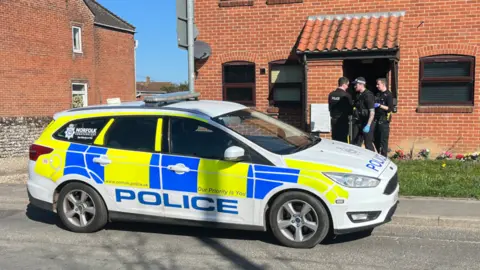 Qays Najm/BBC A marked fluorescent blue and yellow police car parked outside of a property. There are three police officers at the entrance of the building.