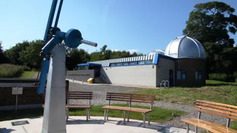 The exterior of an observatory, with wooden benches and a telescope in the foreground. The observatory building comprises a light-coloured brick flat section and a dark-coloured brick curved section which has a silver domed roof on top.