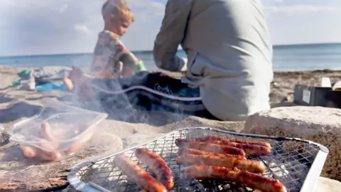 Getty Images A disposable barbecue with sausages being cooked on it. There is a child and dad sitting on a sandy beach in the background.