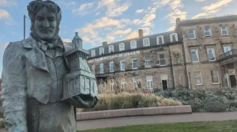 Paul Burnell/BBC Statue of Sir Peter Fleetwood Hesketh with large sideburns, thick wavy hair and the attire of a Victorian gentleman in front of the North Euston Hotel Fleetwood. The hotel is a large sandstone coloured three storey buildin
ng