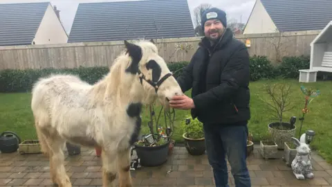 David Lusby A man in a dark jacket, dark trousers and woolly black hat holds a small pony by the reins. He and the horse are stodd in a small garden