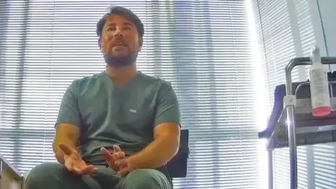 A man wearing medical scrubs is seen talking to someone off-camera.