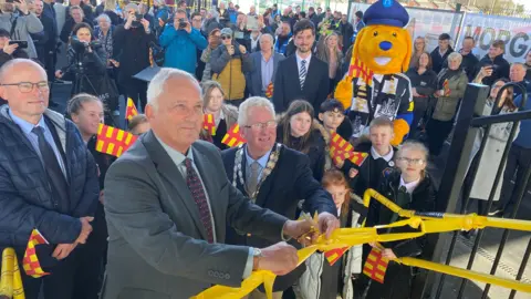 A group of people are watching a politician cutting the ribbon on the steps of a train station. There are children also watching and a large mascot wearing a dog costume and a Newcastle United shirt.