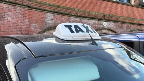 Charlie Stubbs / BBC It shows a white sign, with 'TAXI' printed on in capital black letters. This is on top of a black cab roof and has an aerial going between the left side of the 'X'