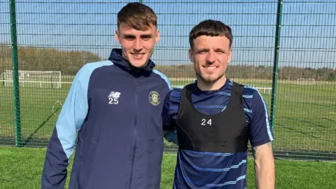 Two young men stand in front of a green mesh fence surrounding a football training ground. One is dressed in a dark blue zip-up training top with the circular crest of Harrogate Town printed on the left breast. The other wears a black training bib with a white "24" numeral on it over a dark blue Harrogate Town strip with light blue stripes.