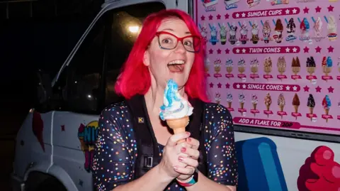 Emma Warley Warley smiles for the camera and is smiling. She is holding an ice cream in a cone in front of a ice cream van. She has red hair, red glasses and wears a polka dot top.