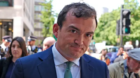 Patrick Spencer leaves a court, dressed in a blue suit and green tie. He is smiling at the cameras. In front of him is a police officer, dressed in uniform. 