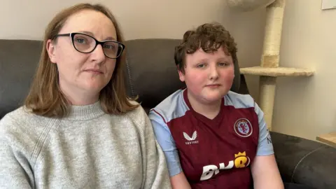 A lady wearing with shoulder length brown hair, glasses and wearing a grey jumper. She is sat next to a young boy with short brown hair and wearing an Aston Villa top. They are sitting on a sofa in a living room looking at the camera.