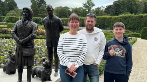 The family of three standing in front of the sculptures.   Cheryl is wearing a white stripy jumper, blue jeans and she is carrying a pink purse and brown pair of sunglasses. She has brown hair and a  fringe. She is smiling.   Gordon is wearing sunglasses on his head. He has brown hair and a subtle beard. He is wearing a white hoody and blue jeans. He is smiling.   Their son is wearing a navy Jack & Jones hoody and black trousers. He has short brown hair and is putting his thumb up, smiling. 